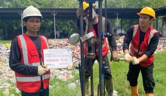 Tiga pekerja teknis Khandaq dengan helm dan rompi keselamatan sedang melakukan uji sondir tanah menggunakan alat mekanis di lokasi proyek Kalideres, memegang papan identifikasi bertuliskan Sondir TS 01.