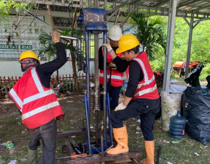 Teknisi geoteknik sedang mengoperasikan dan menekan pipa bor alat uji sondir mekanis ringan secara vertikal di bawah struktur kanopi untuk memetakan karakteristik bawah tanah.