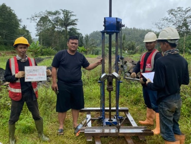 Empat orang anggota tim geoteknik Khandaq berpose bersama peralatan uji sondir di lapangan hijau lokasi proyek PT. Indo Palm Sugar. Tiga pekerja memakai perlengkapan keselamatan penuh, sementara satu orang dengan pakaian kasual menunjuk ke mesin. Pekerja di sebelah kiri memegang papan identifikasi proyek 'SONDIR: TS 01'. Latar belakang menunjukkan perbukitan dan vegetasi hijau di bawah langit mendung