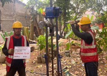 Sebuah deskripsi visual yang terperinci dan berorientasi pada kata kunci untuk aksesibilitas dan SEO: "Sebuah deskripsi visual rinci dua pekerja geoteknik Tim Khandaq sedang mengoperasikan alat uji sondir ringan (Cone Penetration Test/CPT) manual di lokasi proyek Jembatan Krukut, Limo, Depok. Pekerja sebelah kiri memegang papan identifikasi proyek dengan teks jelas 'SONDIR: TS 04' dan logo KHANDAQ. Pekerja sebelah kanan sibuk mengebor dengan alat tersebut. Latar belakang pepohonan hijau di area luar ruangan di Limo di bawah langit mendung."