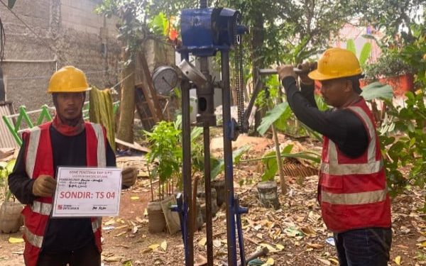 Sebuah deskripsi visual yang terperinci dan berorientasi pada kata kunci untuk aksesibilitas dan SEO: "Sebuah deskripsi visual rinci dua pekerja geoteknik Tim Khandaq sedang mengoperasikan alat uji sondir ringan (Cone Penetration Test/CPT) manual di lokasi proyek Jembatan Krukut, Limo, Depok. Pekerja sebelah kiri memegang papan identifikasi proyek dengan teks jelas 'SONDIR: TS 04' dan logo KHANDAQ. Pekerja sebelah kanan sibuk mengebor dengan alat tersebut. Latar belakang pepohonan hijau di area luar ruangan di Limo di bawah langit mendung."
